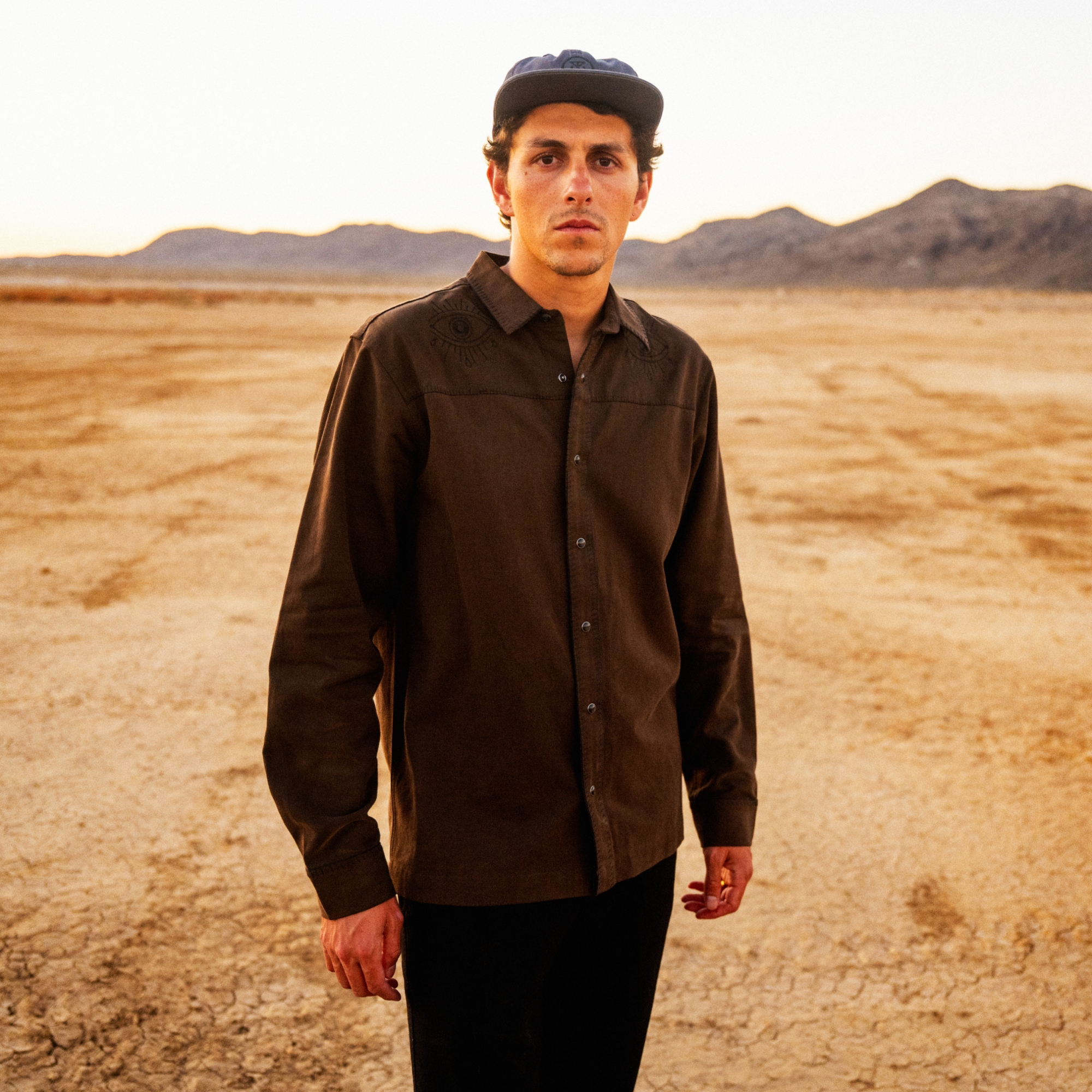 Man wearing brown button-up shirt and black cap standing in desert landscape with mountains in background